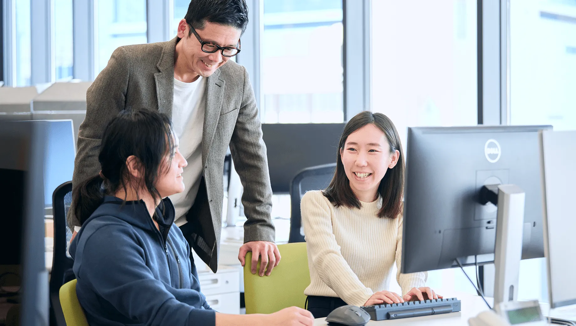 オフィスで笑顔で会話しながらPC画面を見る3人の社員